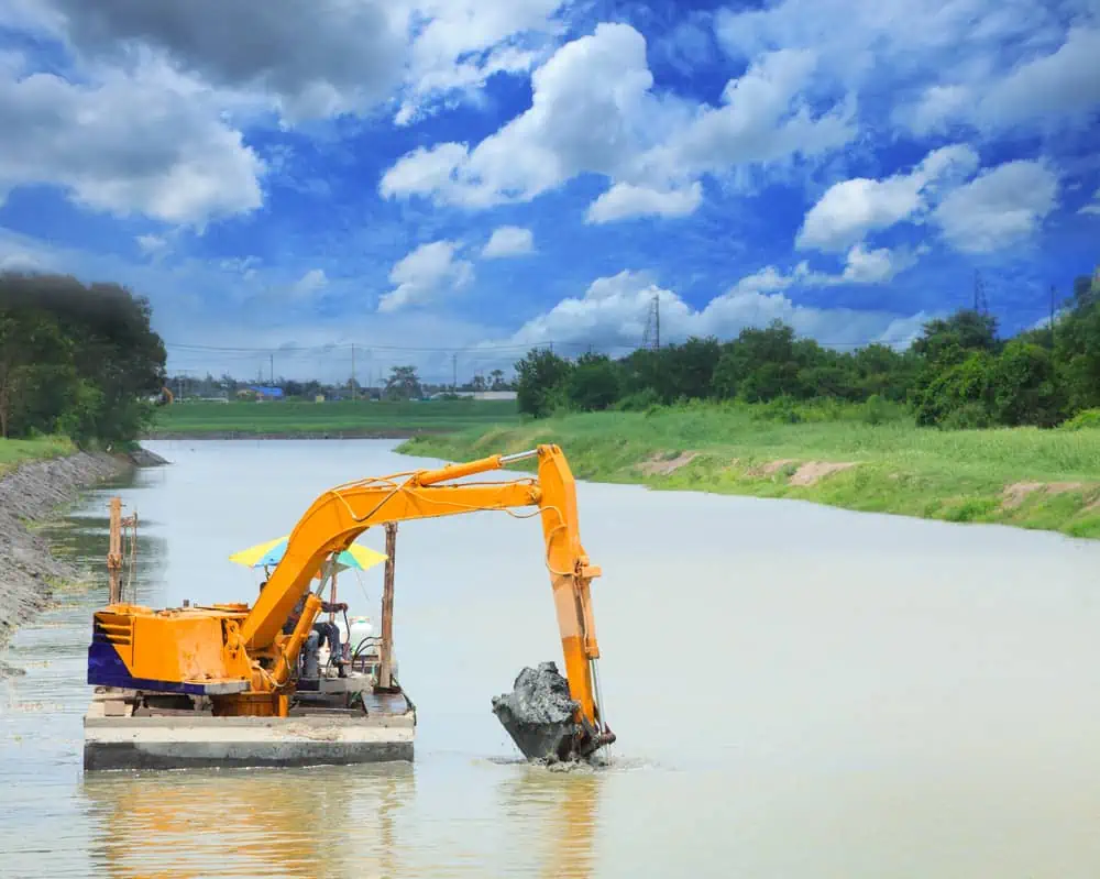An orange excavator is on a boat in a river, performing pond dredging services.