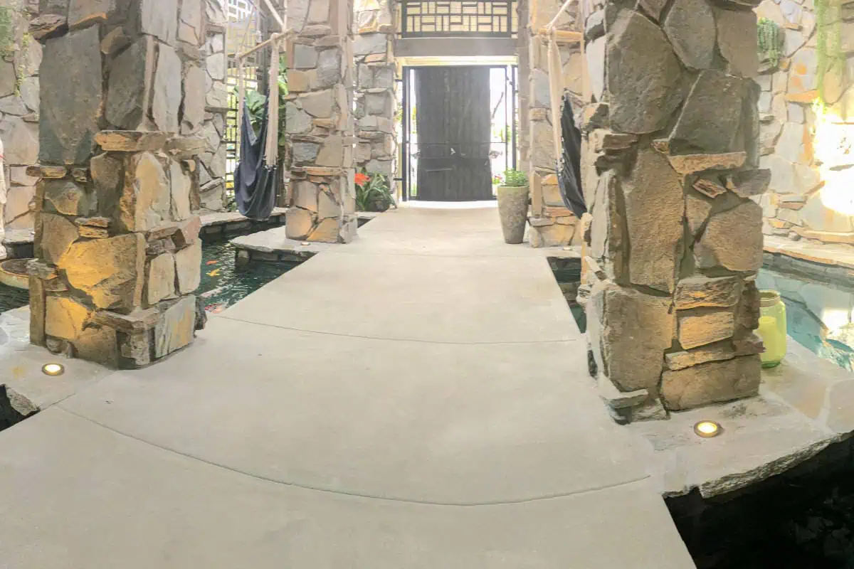 A concrete walkway surrounded by a shallow pool leads to a large black door, with stone columns and planters on either side.