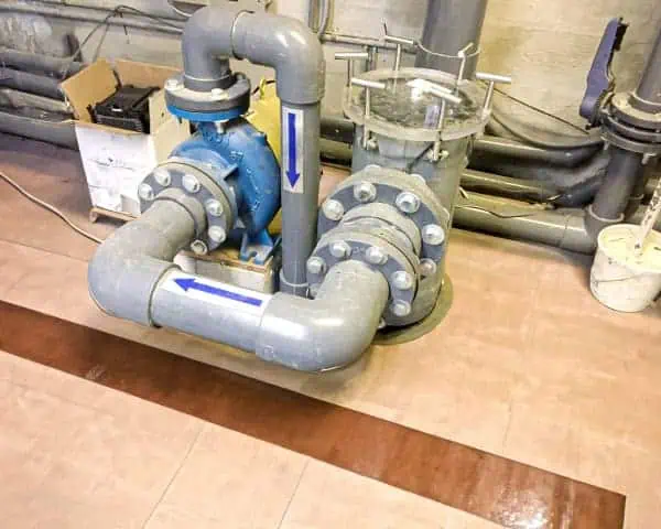 Plumbing system with grey pipes and blue valve, marked with flow direction arrows. It is mounted on a tiled floor amidst other pipes, blending seamlessly into the comprehensive infrastructure typical of Lake Management Services.