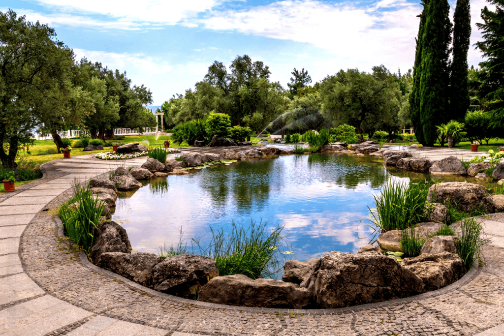 A landscaped garden pond surrounded by large rocks, green plants, and trees, with a stone walkway circling the water under a partly cloudy sky—perfect inspiration for your next Blog Post Template.